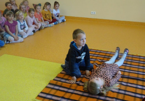 Jedna dziewczynka leży na kocu, chłopiec prezentuje etapy udzielania pierwszej pomocy.